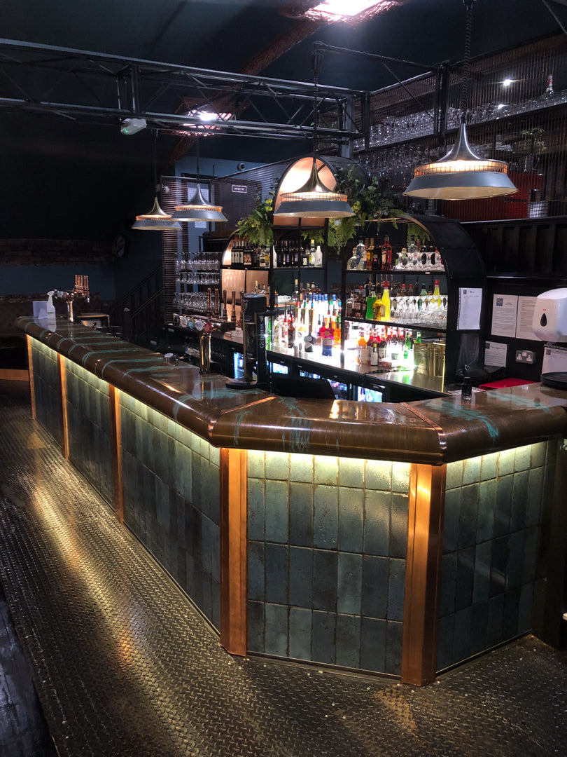 Bespoke copper bar counter with tiled front and illuminated by pendant lights in a dimly lit room.