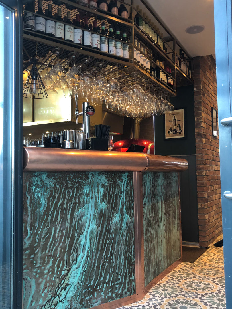 Bespoke copper bar and counter with glasses hanging from a custom steel rack in a modern, handcrafted bar interior.