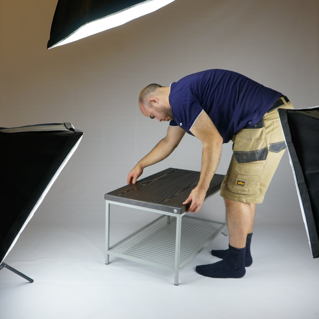 Person assembling a small pine table top on a painted steel frame in a studio setting with lighting equipment.