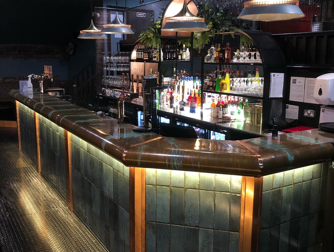 Bar counter with green tiles, wooden accents and a bespoke copper top illuminated by hanging lights.